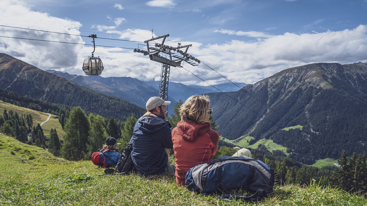 Eine Familie sitzt auf der Bergwiese auf dem Rinerhorn und geniesst die Natur, dahinter schwebt eine Gondel der Rinerhornbahn in Richtung Berg.  | © Davos Klosters Mountains